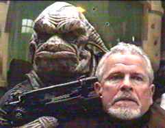 Ein Alien hält dem Priester eine Waffe an die Schläfe