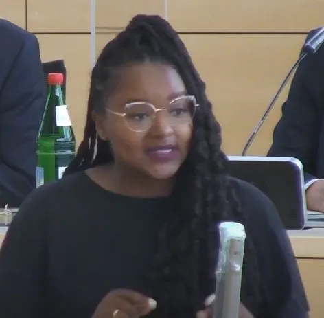 Die dunkelhäutige Politikerin trägt eine Brille, hat lange schwarze Haare und geschminkte Lippen. Vor ihr ein Mikrofon, in das sie spricht.