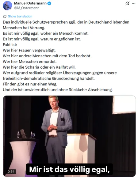 Manuel Ostermann steht in grauem Jacket, weißem Hemd und dunkler Stoffhose mit einem Headset-Mikrofon vor einem Rednerpult.